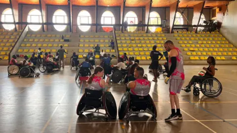 Penrith Pumas Penrith Pumas players and their Ugandan hosts take part in a wheelchair rugby session in a sports hall. The players have their backs to the camera. In the background there are spectator stands with many yellow chairs lining them.