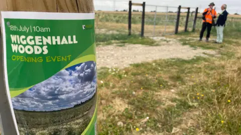 Stephen Huntley/BBC A sign on a wooden post, with two men blurred behind it. One man is wearing a dark jacket and cream trousers, the other man has an orange high-vis jacket and a hat on. There are trees and grass to the right. A field is in the distance.