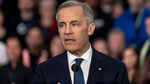 A photo of PM Mark Carney speaking at a podium, with blurred out faces of spectators behind him. He is wearing a black suit and a dark navy polka-dotted tie with a white button up shirt. He has short grey and black hair.