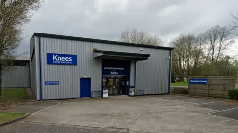 A grey building with a small car park in front. A blue sign says 'Knees home and electrical'