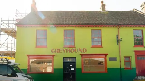 A lime green coloured pub with large red windows and doors. There's a silver works van to the left in front of scaffolding and a skip to the right. 