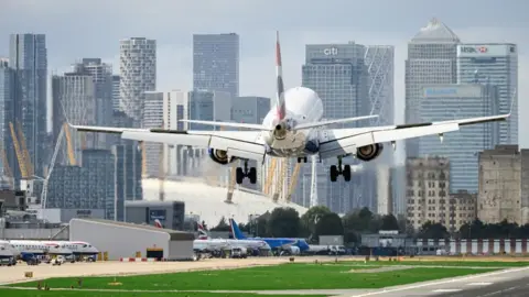  A British Airways aircraft comes in to land at London City Airport. The background has a number of high rise buildings and the O2 arena in Greenwich. 