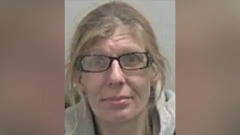 Katie has light brown hair tied up with strands framing her face. She wears black rectangular glasses and a black t-shirt with a grey hoodie