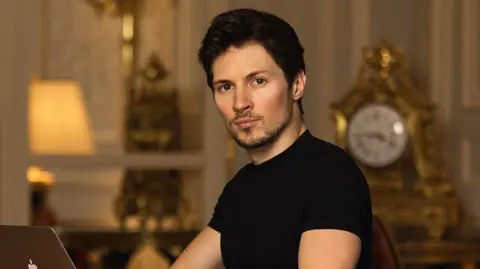 Pavel Durov/Instagram Tech billionaire Pavel Durov looks to the camera as he types on an Apple laptop. He is wearing a black t-shirt. There is an ornate clock in the background.