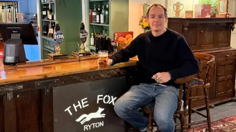 A man (Steve Lee) sat at a barstool, wearing light blue jeans and a black woollen sweatshirt. He's holding a half-full pint glass in his right hand and is looking towards the camera and smiling slightly. The bar is dark wood, with a sign on its base that reads 'The Fox, Ryton' in white with an illustration of a running fox. The walls of the pub are painted light green and the floor is black tiling with a red patterned rug. 
