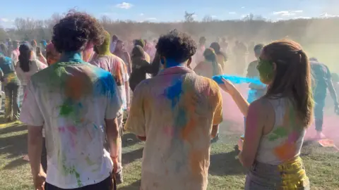 Jenny Kirk/BBC Three people are standing with their backs towards the camera. They are all wearing white T-shirts and have lots of coloured power on the back. In front of them there are lots of people throwing colourful powders at each other. 