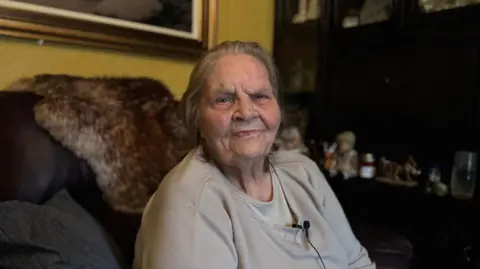 Annie Sweeney sitting on a chair in a living room.  She has straight, grey hair swept back from her face and is wearing a beige jumper and T-shirt. 