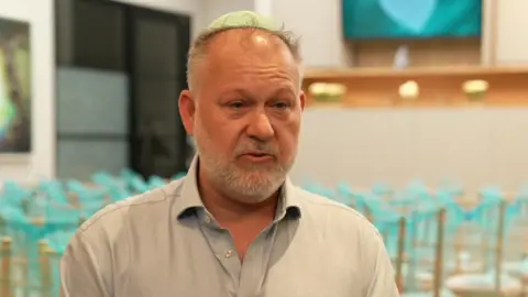 Adam Ziff wears a light green kippah and speaks while looking slightly to the side in a room with chairs covered in blue ribbons.