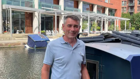 Sofía Luis-Hobbs/BBC Ken Marshall, a middle-aged man wearing a blue polo shirt standing next to a narrow boat. He has short grey hair and is smiling in the photo.