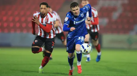 A football player in a red and white jersey is running beside another player, who is in a dark blue jersey. There are two other players in red and white jerseys behind them.