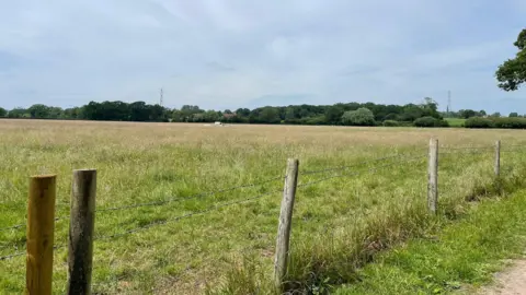 A large open field in Pewsham, Chippenham with trees and hedgerows in the background and fence posts with barbed wire in the foreground.