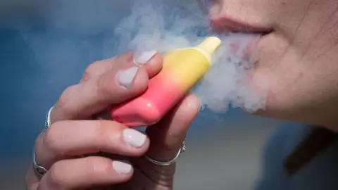 A woman holds a yellow and red coloured vape to her mouth with a cloud of vapour surrounding it.