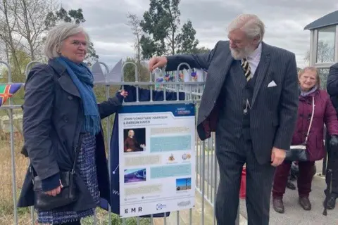 A woman and a man remove a blue curtain covering an information board on a railway station platform. The woman has grey hair and wears a blue coat and matching dress and scarf. The man as white hair, a full beard and wears a three-piece grey pin-striped suit. The board, primarily coloured white and blue, features text and images, including a picture of a man wearing 17th Century clothing and the words: "John 'Longitude' Harrison & Barrow Haven".