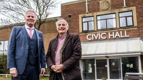 Stuart Boulton Glenn McCrory, from Lamplight Arts Centre Ltd, and councillor James Rowlandson at the former Civic Hall in Stanley.  Mr McCrory is wearing a blue three-piece suit and a pink tie. Next to him, on the right is Rowlandson wearing a thick, brown coat. The entrance to the civic hall is behind them.