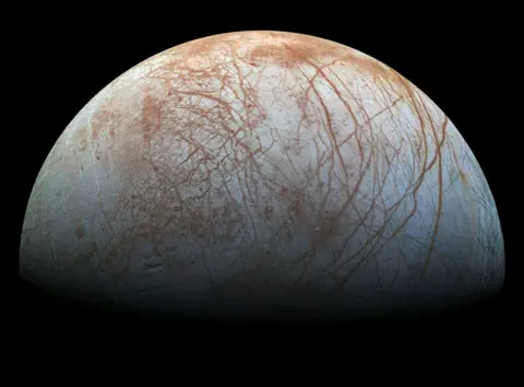 La luna helada de Júpiter de la NASA, Europa, ocupa un lugar destacado en esta vista de imágenes tomadas por la nave espacial Galileo de la NASA. La luna aparece plateada con grietas anaranjadas en su superficie.