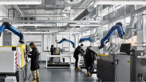 UniversityofManchester Lab workers wear black coats and goggles as they work in a lab at the Graphene Engineering Innovation Centre. Blue suction tubes can be seen overhead with various other pieces of scientific equipment seen. 