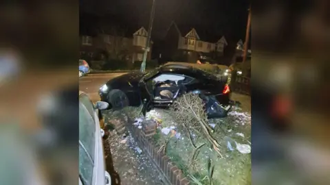 Surrey Police A black car with its side door open is partly in a front garden, where a tree is seen damaged alongside it and a part of a low wall has been knocked down. It is dark and there are semi detached houses seen in the background