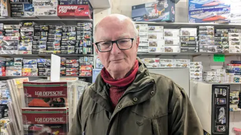 A white man with black glasses and white hair, wearing a dark green jacket and red jumper. Behind him are stacks of model making kits. 