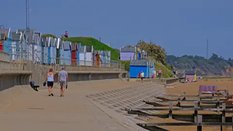 Squiz/BBC Weather Watchers A coastal area on a sunny day. People are walking along a promenade. There are some beach huts above them.