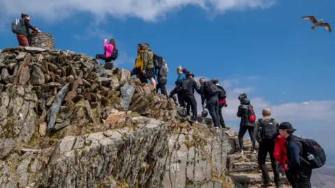 Getty Images People wearing coats and backpacks queue to reach the top of a mountain, against the backdrop of a blue sky.