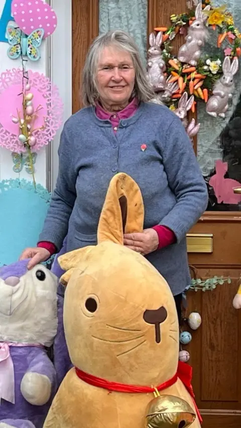 A woman standing in front of a brick house doorway decorated for Easter with colourful eggs, butterflies, hanging baskets and a “Happy Easter” banner, surrounded by plush bunnies and other festive ornaments.
