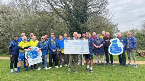 A group of people from the walking group wearing, mainly, blue tops and holding up a large cardboard cheque. There are trees and hedges behind them.