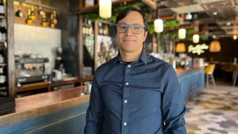 Rogerio looks at the camers with the bar in the background. He is wearing a dark blue shirt and has glasses on with a dark frame. His black hair covers some of his forehead.