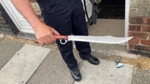 A man wearing black trousers and boots, you can only see bottom-half of his body, as he holds a large machete style knife, with a brown handle. The man is standing on a pavement outside a terraced house.