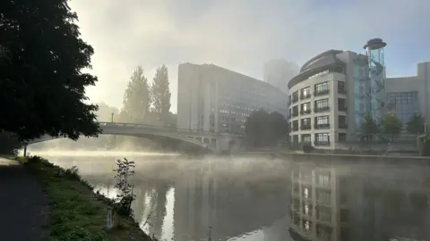 EmmaH Mist rolls over a river in a built-up suburban area. A bridge crosses the river with apartment blocks on one side. Trees can be seen on the other side under a murky sky, although part of it is blue.