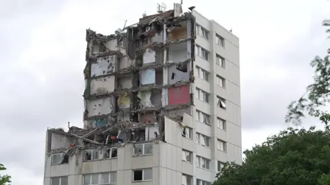 A tower block that is part demolished. Rows of flats have been taken out. It is standing against a grey background.