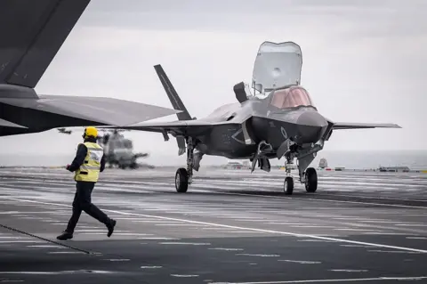 Getty Images A member of the flight deck crew runs past as an F-35B, the STOVL (short takeoff and vertical landing) variant of the Lockheed Martin F-35, takes off from the flight deck of HMS Prince of Wales (R09), a Queen Elizabeth-class aircraft carrier