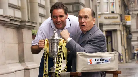 SNS Ally McCoist and Robert Duvall hold the Scottish Cup. Duvall is in a director's chair while McCoist is leaning forward in a white T-shirt.
