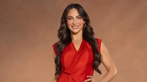 Paige Chohan, who is wearing a red dress and gold hooped earrings, is smiling at the camera in this promotional image for the new series. 