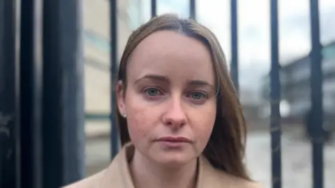 A woman with long light brown hair, wearing a beige coat. There is a grey wall behind her and black railings