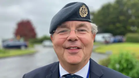 Derek Coad smiling at the camera. He is wearing a beret and a black suit jacket, with a white shirt. 