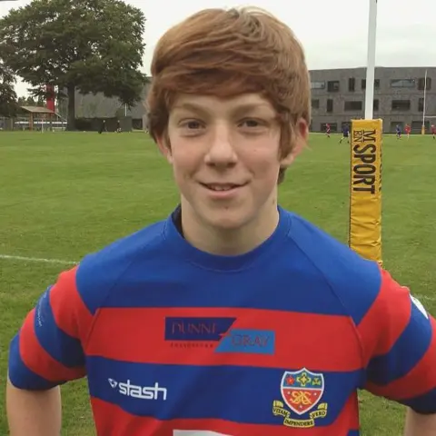 CIARAN A young Ciaran Booth stands on a rugby pitch wearing a red and blue striped rugby shirt.