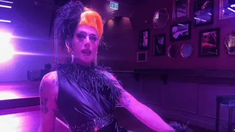 Drag queen in a wig sitting on the stage of the Black Cap pub where there is a purple light shining on her, she wears a one-shoulder dress/top with ostrich feathers decorating the diagonal line which gathers at one shoulder. Her hair is up and she has full make up, earrings on and tattoos on her upper shoulders. In the background is the stage and on the right, the wall with various framed photos on it.