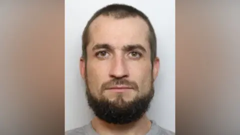 A close-up mugshot of a man with brown eyes, beard, eyesbrows and hair. He is looking directly at the camera and is in front of a light grey background
