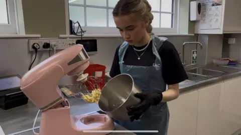 A teenage girl with blonde hair, wearing a blue plastic apron is using a pink cake mixer on a work bench.
She is holding the bowl