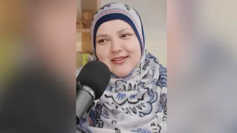 A woman in a blue and pink floral headscarf smiles into a microphone.