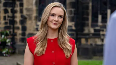 LDRS A person with long, light-coloured hair stands outdoors in front of a stone building with dark window frames. The person is wearing a bright red sleeveless top with large buttons down the front. The background appears slightly out of focus, showing textured stone walls and greenery at ground level. The image is a close-up portrait, with soft natural lighting highlighting the subject against the darker architectural backdrop.