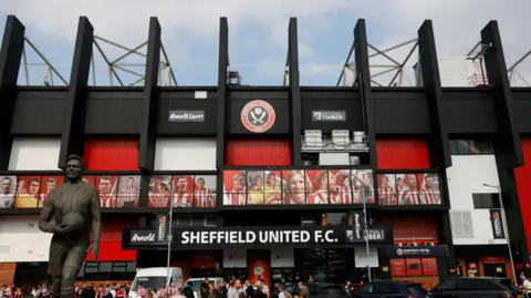 PA Media Fans can be seen in front of the entrance to Bramall Lane, which features a sign that reads "Sheffield United FC". A bronze statue of a man can be seen on the left. The stadium's colour scheme is white and red.