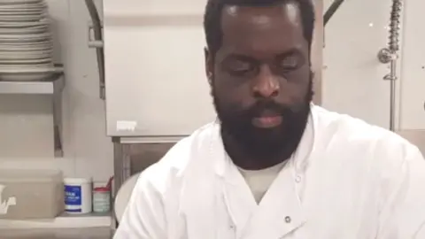 Jomi Denedo working in a kitchen, he is wearing a white chefs suit, and black gloves, he is cleaning a pan. Kitchen items are around him including paper towels, a sink and plates and cups on a shelf to the left. He is looking down, and has a beard.