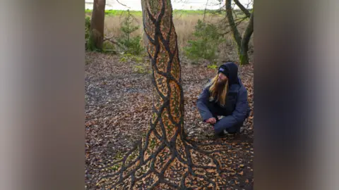 Jon Foreman/Sculpttheworld A man kneeling by a tree he has covered in autumnal yellow and orange leaves in an artistic pattern.