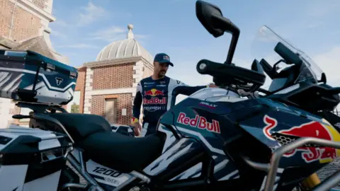 Two-time Dakar Rally Champion Sam Sunderland by his Triumph Tiger 1200 Rally Explorer motorbike. He is wearing his racing outfit. It is a sunny day.