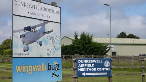 A sign showing a light aircraft with Dunkeswell Aerodrome written above. There is also a navy blue sign mounted to a wooden fence that reads Dunkeswell Airfield Heritage Centre.