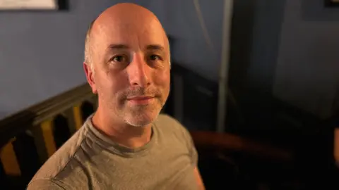 A man looks a the camera with a slight smile on his face. He has a grey t-shirt on and grey stubble. Behind him is a blue wall. 