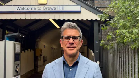 A middle-aged man with a grey quiff and black glasses looks into the camera. He is wearing a light blue jacket