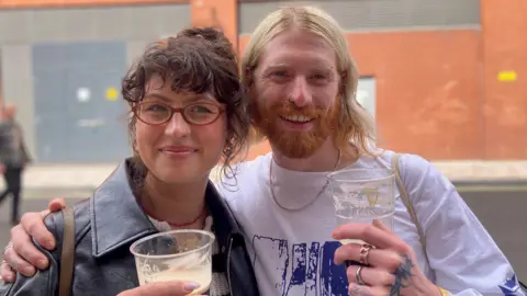 Two people smiling at the camera. On the left is a woman with dark curly hair and glasses. She is wearing a black leather jacket and is holding a plastic cup of Guinness. On the right is a man with long blond hair and an auburn beard. He is wearing a white t-shirt and is holding a plastic cup of Guinness. 
