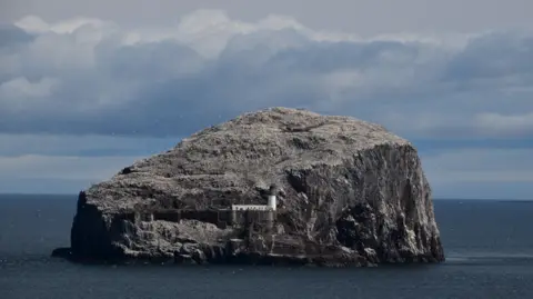 Lynsey Bell A large grey rock in a body of water. There are dramatic clouds and birds flying close to the rock, and a white lighthouse in the middle of the scene.